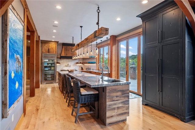 kitchen with a breakfast bar, dark stone countertops, light hardwood / wood-style flooring, a kitchen island with sink, and premium range hood