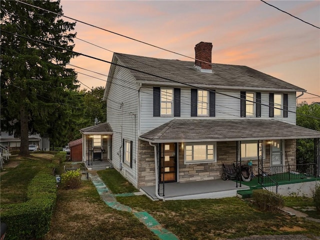 view of front facade with a chimney, a shingled roof, a porch, a lawn, and stone siding