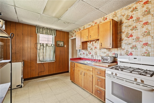 kitchen with brown cabinets, white gas stove, a paneled ceiling, wood walls, and a sink