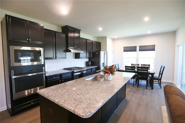 kitchen with hardwood / wood-style flooring, appliances with stainless steel finishes, light stone countertops, sink, and an island with sink