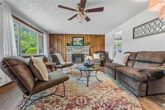 living room featuring a stone fireplace, wood walls, wood-type flooring, ceiling fan, and a textured ceiling