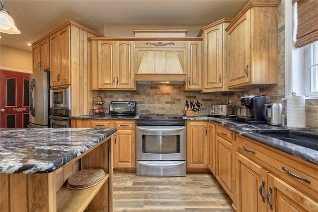 kitchen featuring dark stone countertops, backsplash, light wood-type flooring, stainless steel appliances, and premium range hood