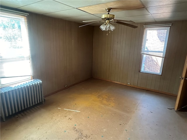 unfurnished room with wood walls, ceiling fan, radiator heating unit, and a drop ceiling