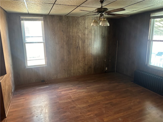 empty room featuring dark hardwood / wood-style floors, a drop ceiling, wood walls, ceiling fan, and radiator