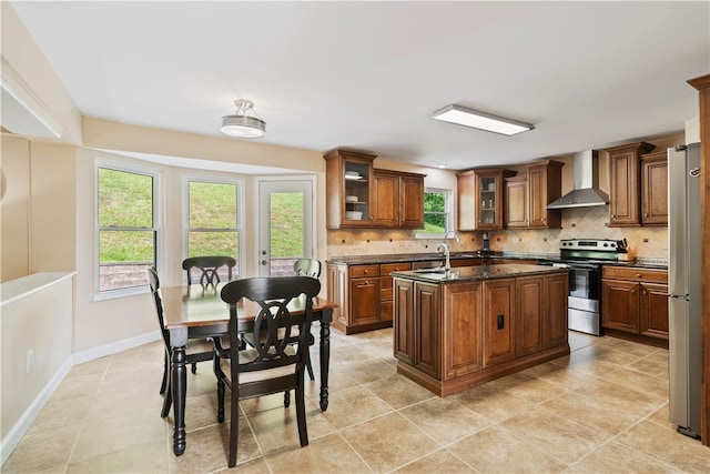 kitchen featuring tasteful backsplash, wall chimney range hood, stainless steel appliances, dark stone countertops, and a kitchen island with sink