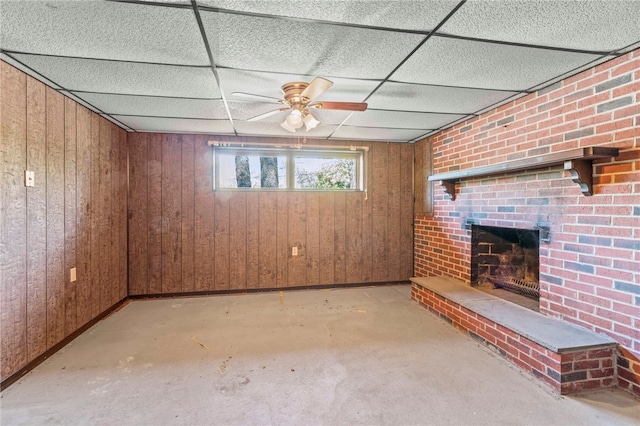 basement featuring a paneled ceiling, ceiling fan, wooden walls, and a brick fireplace