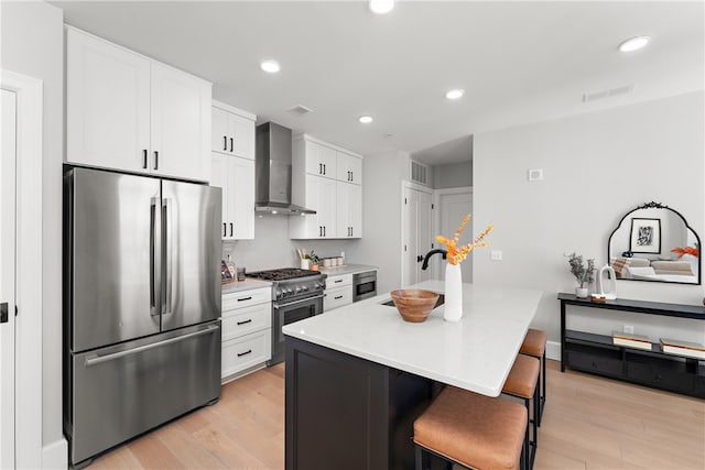 kitchen featuring appliances with stainless steel finishes, a kitchen island with sink, white cabinetry, wall chimney range hood, and light wood-type flooring