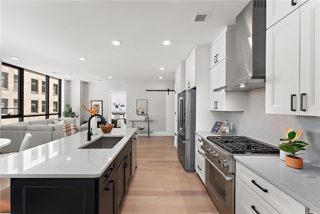 kitchen featuring a barn door, light wood-type flooring, stainless steel appliances, wall chimney exhaust hood, and a center island with sink