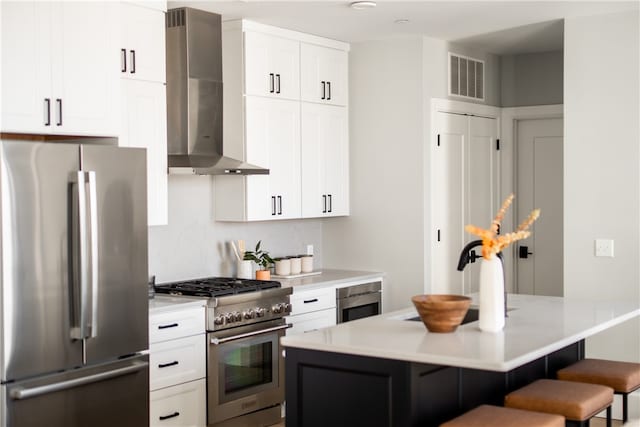 kitchen with a kitchen island, a breakfast bar, appliances with stainless steel finishes, white cabinets, and wall chimney range hood