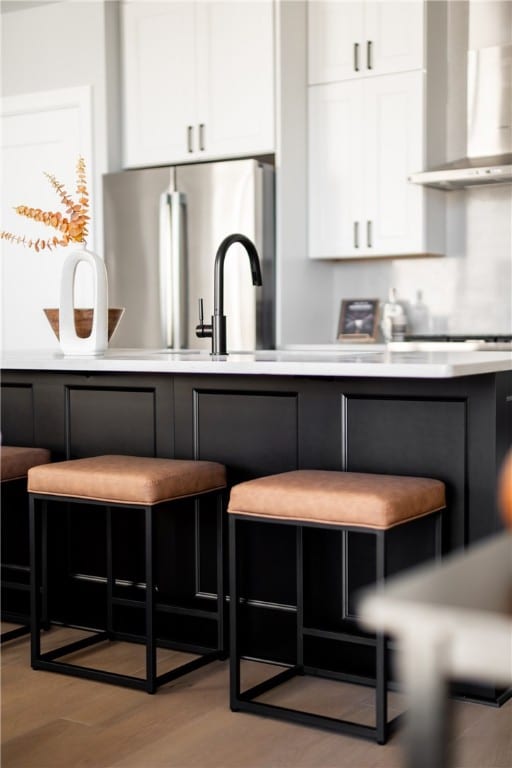 kitchen featuring light hardwood / wood-style floors, stainless steel refrigerator, white cabinetry, wall chimney range hood, and a breakfast bar