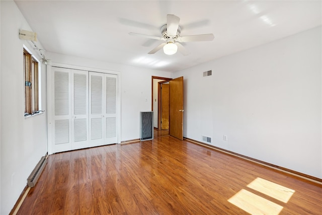 unfurnished bedroom with baseboard heating, wood-type flooring, ceiling fan, and a closet