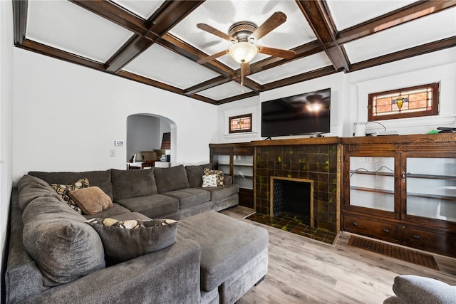 living room with a tiled fireplace, ceiling fan, light wood-type flooring, beamed ceiling, and coffered ceiling