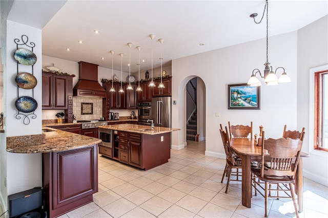 kitchen featuring an island with sink, custom exhaust hood, an inviting chandelier, stainless steel appliances, and decorative light fixtures