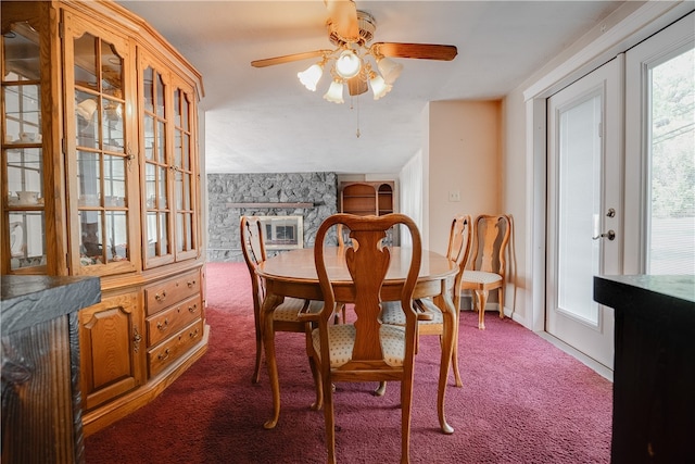 dining area with ceiling fan, a stone fireplace, and dark colored carpet