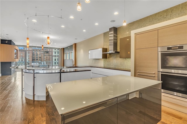 kitchen with double oven, hanging light fixtures, wall chimney range hood, and dark hardwood / wood-style flooring