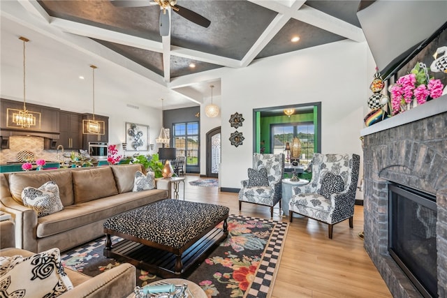 living room with ceiling fan with notable chandelier, beam ceiling, light hardwood / wood-style flooring, and a stone fireplace