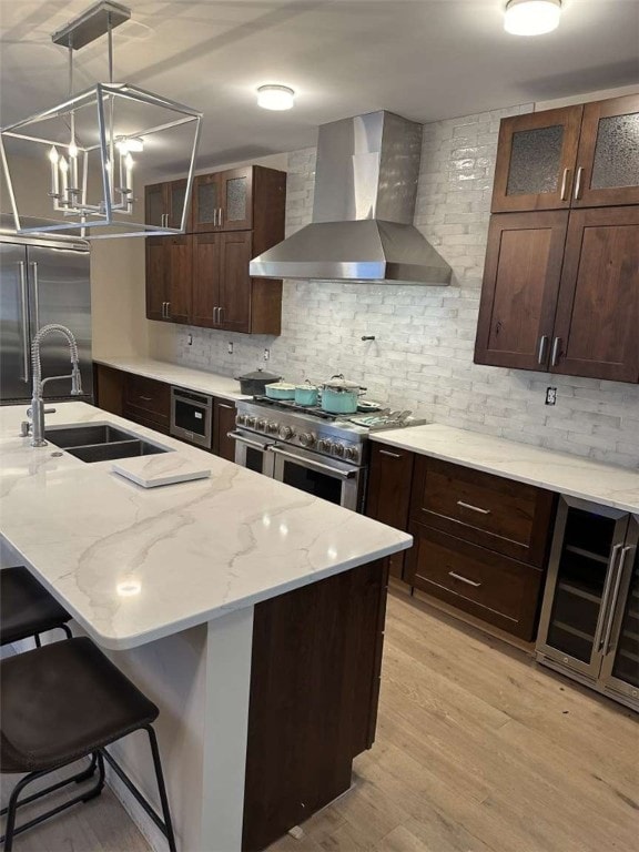 kitchen featuring high end appliances, beverage cooler, wall chimney range hood, light wood-type flooring, and a kitchen bar