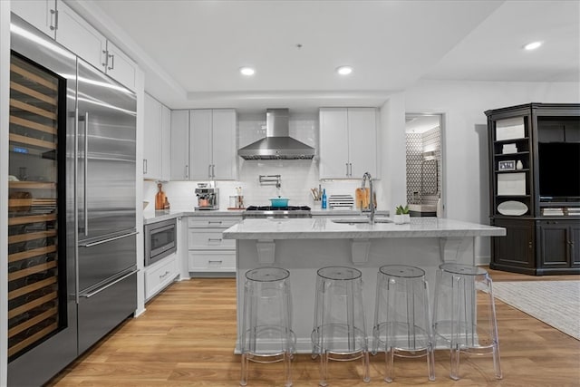 kitchen with white cabinets, a breakfast bar area, built in appliances, a kitchen island with sink, and wall chimney range hood