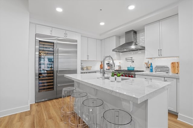 kitchen featuring a center island with sink, wall chimney range hood, white cabinetry, appliances with stainless steel finishes, and a breakfast bar