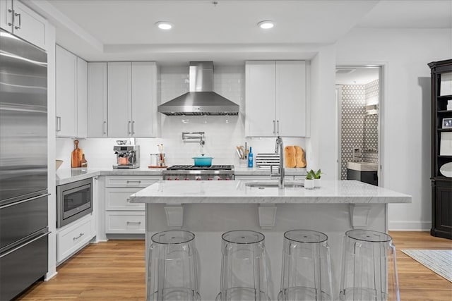 kitchen with light hardwood / wood-style floors, white cabinetry, wall chimney exhaust hood, built in appliances, and sink