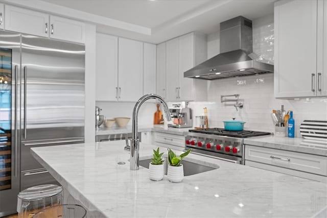 kitchen with decorative backsplash, light stone counters, white cabinets, wall chimney exhaust hood, and premium appliances