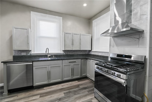 kitchen with gray cabinetry, dark stone countertops, wall chimney exhaust hood, stainless steel appliances, and hardwood / wood-style floors