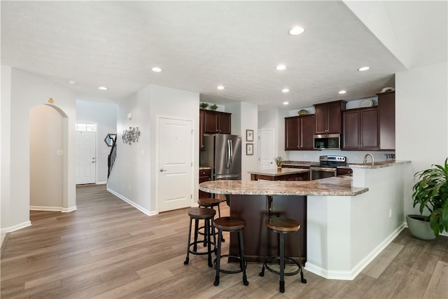 kitchen with a kitchen breakfast bar, light wood-type flooring, appliances with stainless steel finishes, dark brown cabinets, and light stone counters