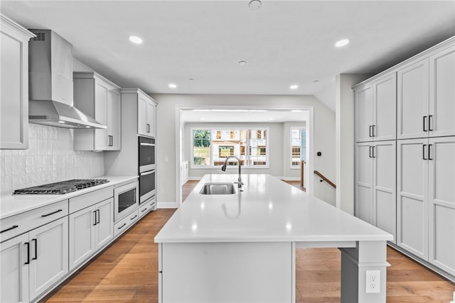 kitchen featuring light wood-type flooring, sink, wall chimney exhaust hood, stainless steel appliances, and a center island with sink