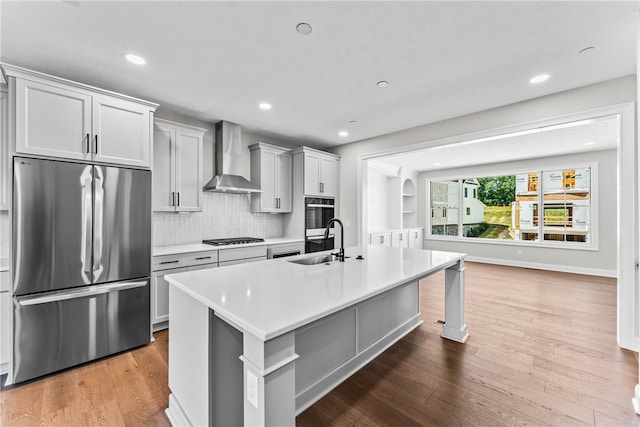 kitchen featuring sink, an island with sink, wall chimney range hood, appliances with stainless steel finishes, and hardwood / wood-style floors