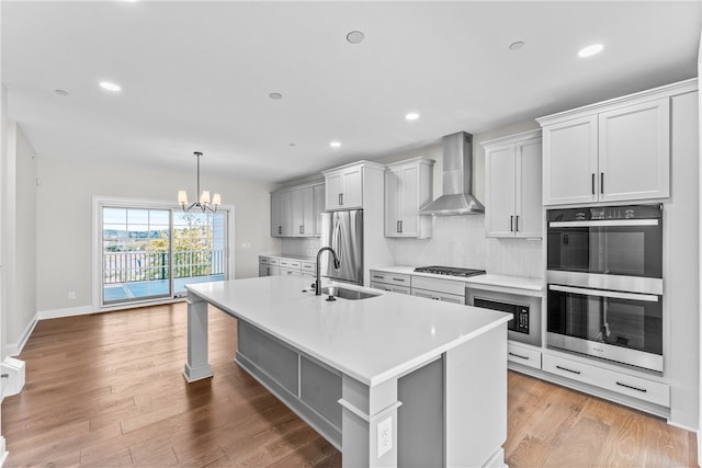 kitchen featuring an island with sink, wall chimney exhaust hood, stainless steel appliances, light wood-type flooring, and sink