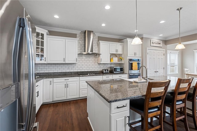 kitchen with a kitchen island with sink, stainless steel appliances, wall chimney range hood, white cabinetry, and decorative light fixtures