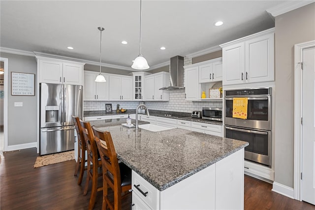 kitchen with white cabinets, an island with sink, hanging light fixtures, wall chimney exhaust hood, and stainless steel appliances