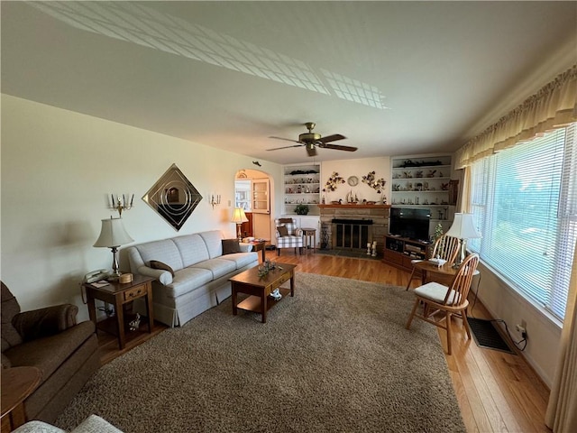 living area featuring a glass covered fireplace, built in features, a ceiling fan, visible vents, and wood finished floors