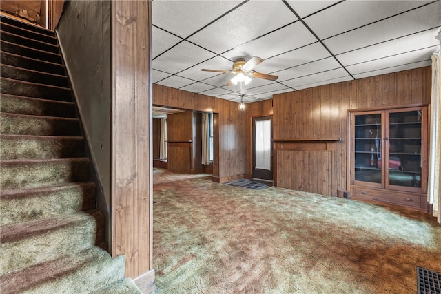 unfurnished living room with ceiling fan, a paneled ceiling, wood walls, and carpet