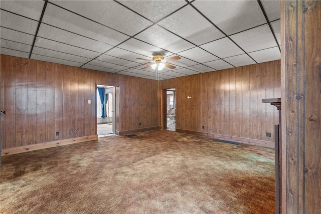 carpeted spare room with a drop ceiling, wooden walls, and ceiling fan