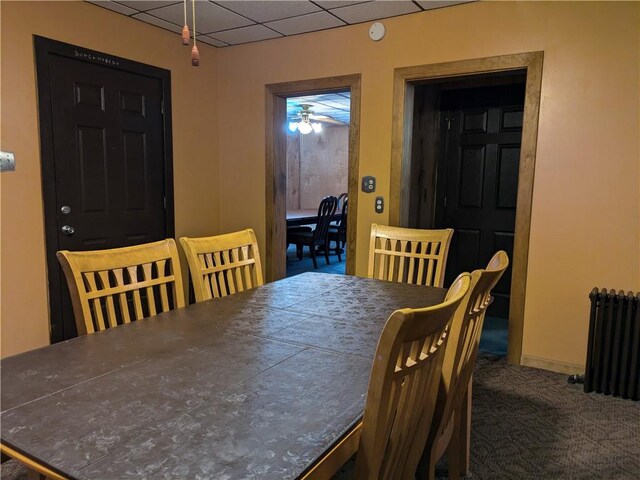 carpeted dining space with a drop ceiling, ceiling fan, and radiator heating unit