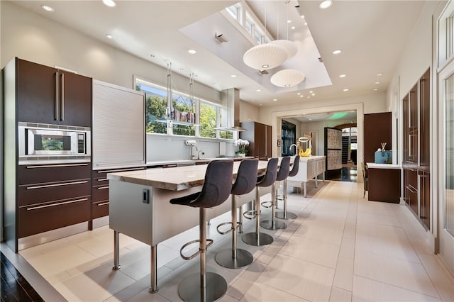 kitchen with light stone counters, island exhaust hood, a kitchen bar, hanging light fixtures, and a raised ceiling