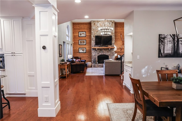 dining space featuring wooden walls, dark wood-type flooring, a stone fireplace, and a chandelier