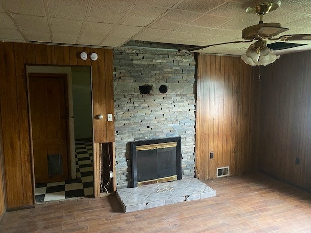 unfurnished living room featuring wood walls, a drop ceiling, a stone fireplace, ceiling fan, and light hardwood / wood-style floors