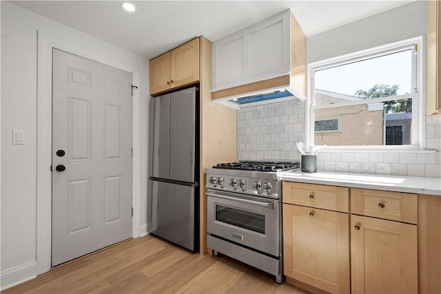 kitchen with light brown cabinets, premium range hood, backsplash, light wood-type flooring, and appliances with stainless steel finishes