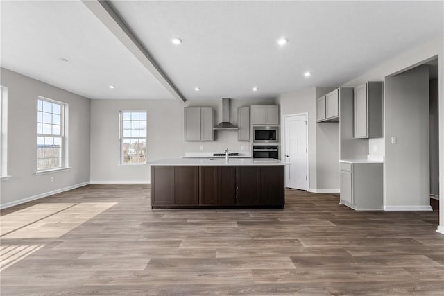 kitchen with wall chimney exhaust hood, stainless steel appliances, an island with sink, wood-type flooring, and gray cabinets