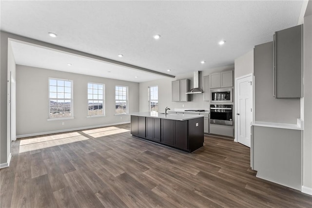 kitchen with appliances with stainless steel finishes, gray cabinetry, a kitchen island with sink, dark wood-type flooring, and wall chimney range hood