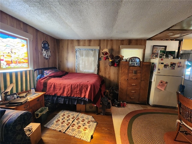 bedroom with a textured ceiling, wood finished floors, freestanding refrigerator, and wooden walls