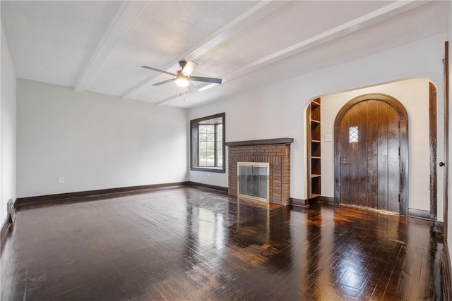 unfurnished living room featuring beam ceiling, dark wood-type flooring, a brick fireplace, and ceiling fan