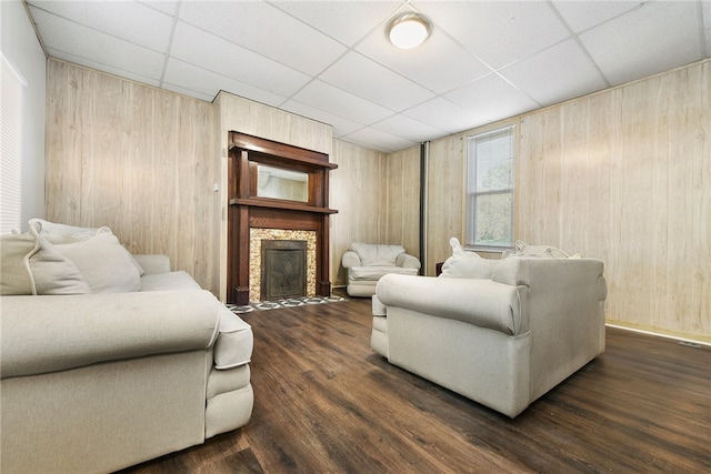 living room with a stone fireplace, wooden walls, a drop ceiling, and dark hardwood / wood-style floors