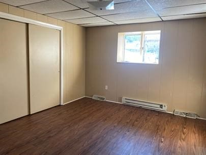 unfurnished bedroom with a drop ceiling, a closet, dark wood-type flooring, and a baseboard heating unit