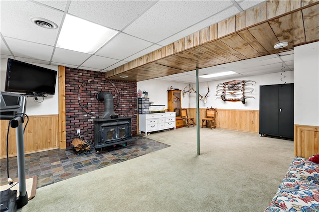 basement with wooden walls, a drop ceiling, carpet flooring, and a wood stove