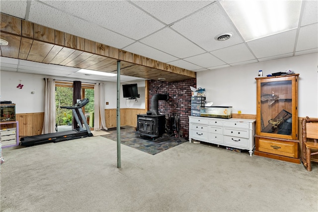 basement featuring carpet floors, wood walls, a wood stove, and a paneled ceiling