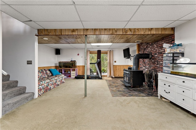 basement with carpet, a drop ceiling, wooden walls, and a wood stove