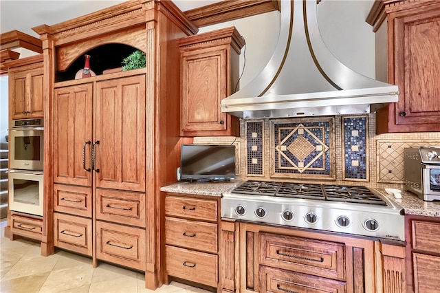 kitchen featuring light stone counters, stainless steel appliances, light tile patterned flooring, wall chimney range hood, and backsplash